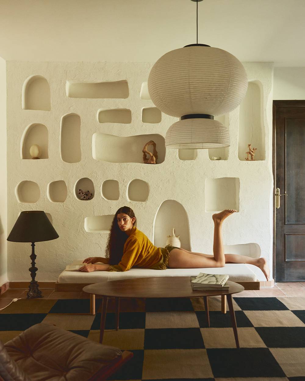 Picture of a woman laying on her stomach on a white bench. The woman is wearing a yellow and green short Terry dress. In the background you can see a bookshelf, overhead there's a ricepaper lamp and underneath the bench there's a checked rug. 