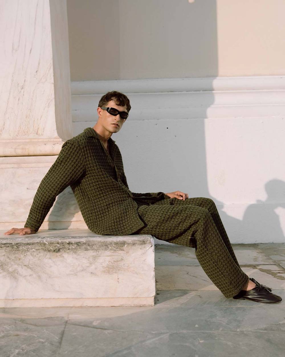 A cropped image of a man leaning against a dark wooden wall. He is wearing a light green and cream Terry cotton pants in a banana leaf design.