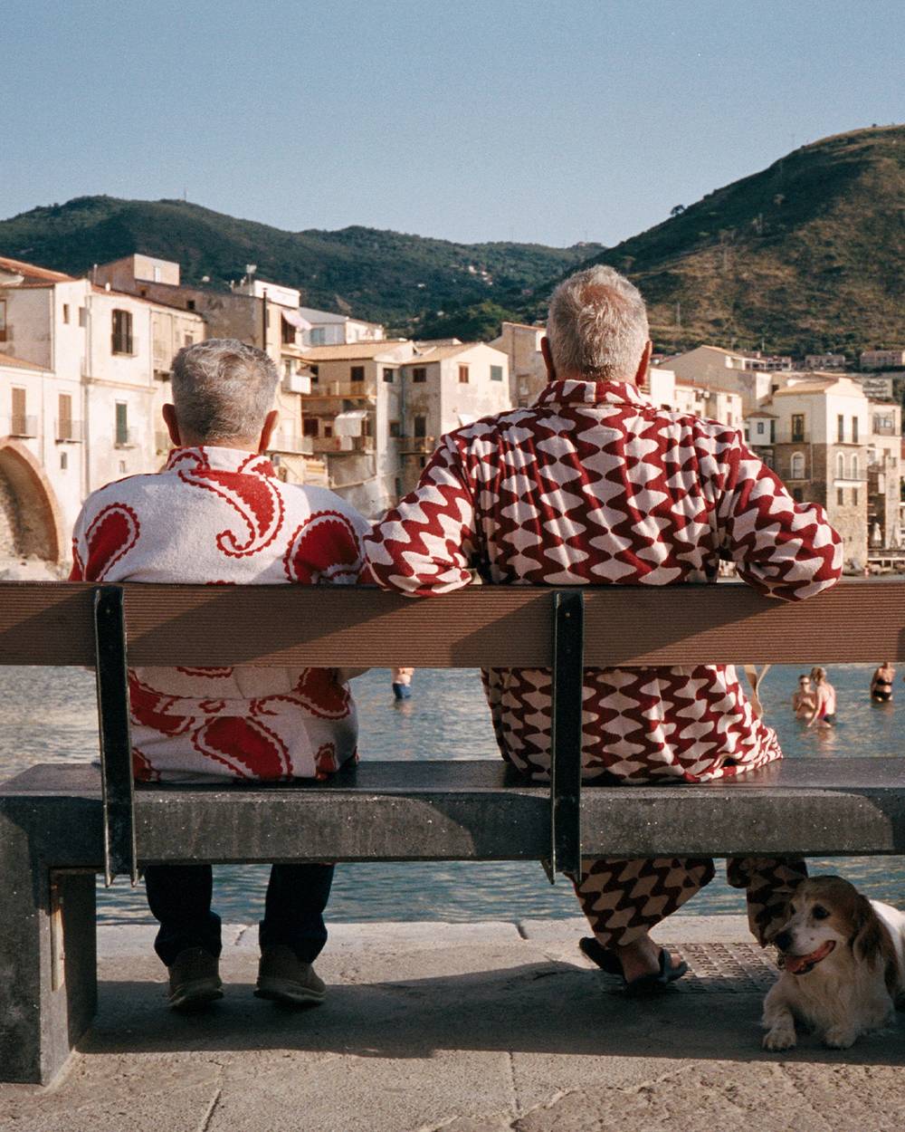 A horizontal picture shot from behind of two elderly men sitting on a bench. The man on the left is wearing a red and white paisley bathrobe and the man on the right a red and white wavy pattern bathrobe. The two men look out over a bay on the Mediterranean where people are swimming and enjoying the sun weather.