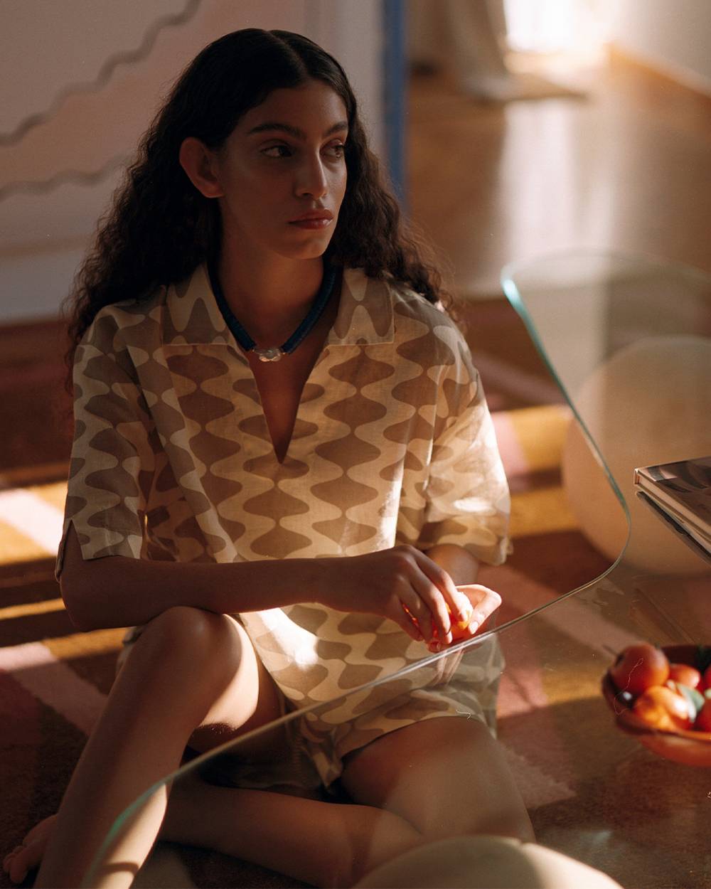 A woman sitting on the floor next to a glass-table with the light seeping in creating a shadows all around. She is wearing a cream and sand printed collared shirt and matching shorts.