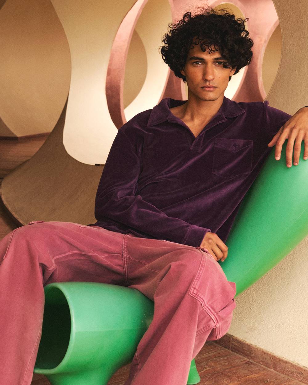A dark and curly haired man is sitting on a bright green chair in a hallway. He is wearing a long sleeved deep purple velour shirt and washed red cargo pants.