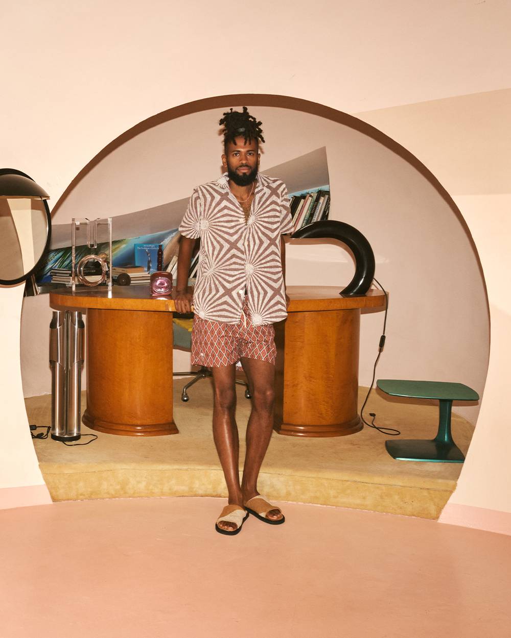 A man standing in an arched opening to an office, leaning on the desk. He is wearing a brown and white printed Terry shirt, a pair of rusty red diamond printed swim shorts and a pair of caramel brown and cram printed Terry slippers. The room he's standing in is a light shade of pink.