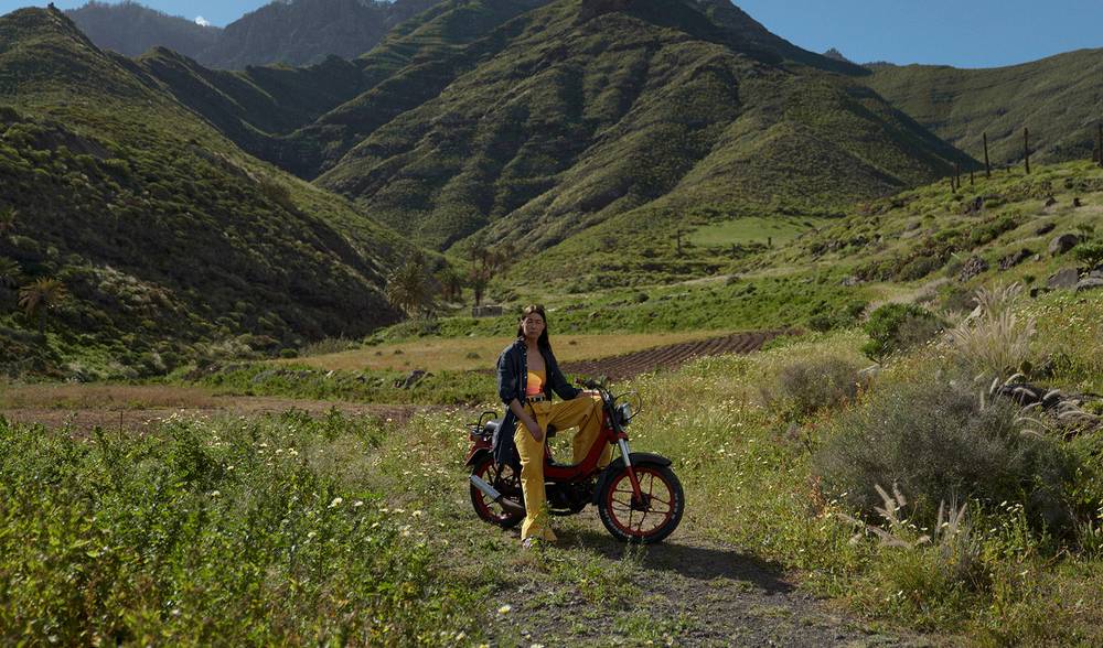 Set high up in the green mountains on a field a woman is half sitting on a red moped. She is wearing yellow pants, a yellow and red bathinsuit as a top and a navy linen kaftan.  