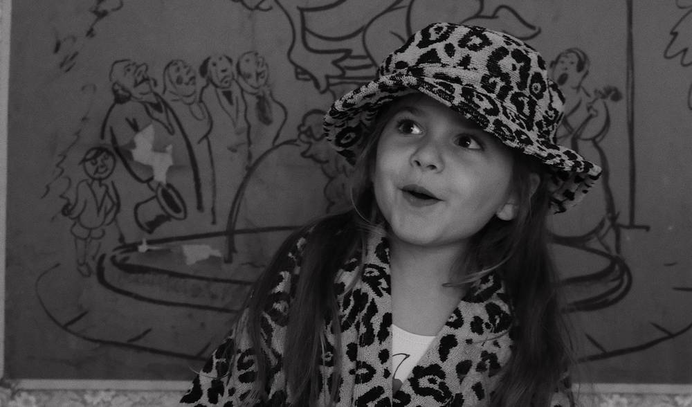 A black and white photo of a small girl with long hair. She is wearing a leopard printed bathrobe and a matching bucket hat.