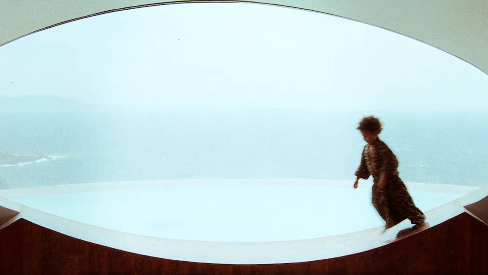 A wide set photo of a kid running on a deep windowsill of a round window wearing a leopard printed bathrobe. The background is light blue and you can almost see the horizon of the ocean.
