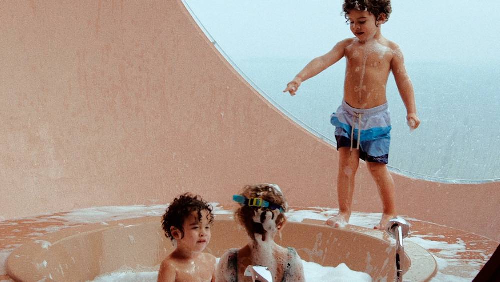 Three boys are playing in a sunken bathtub filled with bubbles. One is standing on the ledge of it and pointing at the other kids, he is wearing a pair of blue monochromatic swim shorts. The other two are sitting down and one is wearing swim googles.  