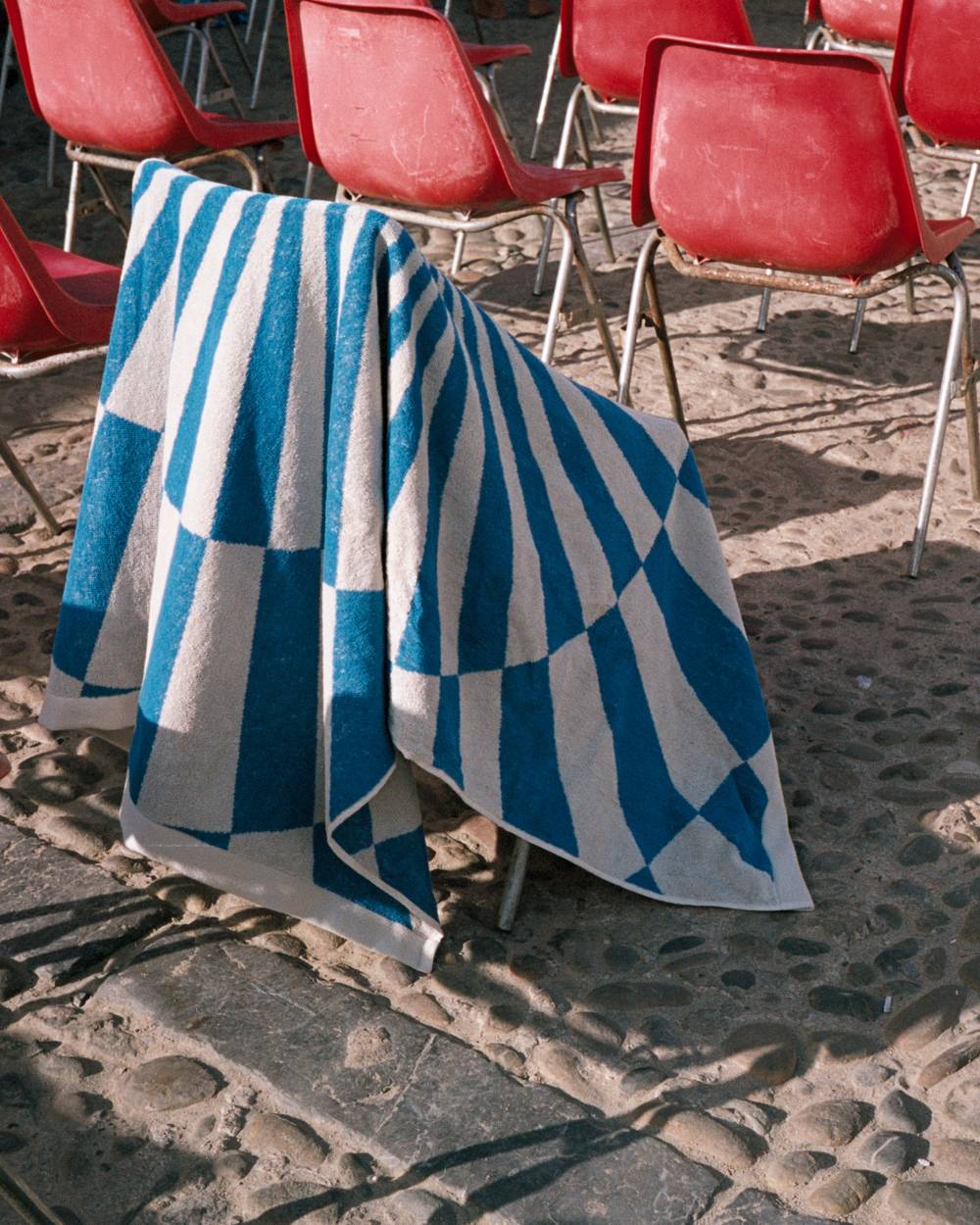 A blue and white towel hanging on a chair on a cobblestone beach. In the background you can see similar red chairs standing around.