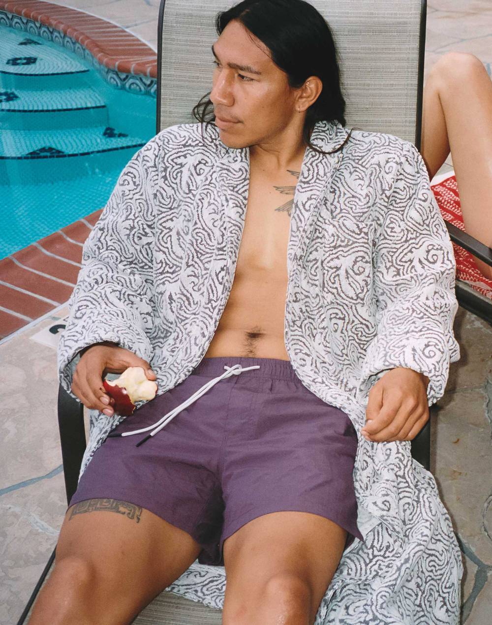 A cropped image of a young man wearing a pair of navy swim shorts with a yellow sun print. He has a burgundy and white printed towel thrown on his shoulder as well. Blurred in the front of the picture there is two men sitting one is wearing black and the other one is wearing a purple velour shirt.  