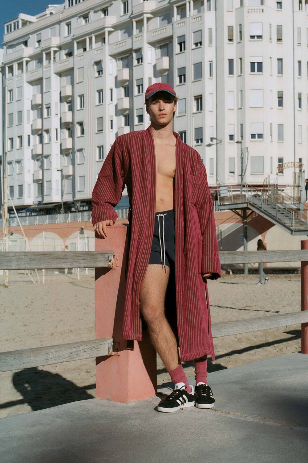 A cropped image of a young man wearing a pair of navy swim shorts with a yellow sun print. He has a burgundy and white printed towel thrown on his shoulder as well. Blurred in the front of the picture there is two men sitting one is wearing black and the other one is wearing a purple velour shirt.  