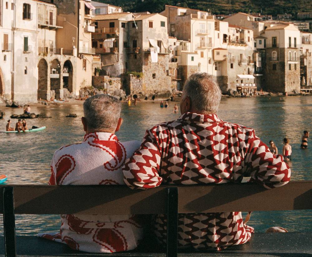 A horizontal picture shot from behind of two elderly men sitting on a bench. The man on the left is wearing a red and white paisley bathrobe and the man on the right a red and white wavy pattern bathrobe. The two men look out over a bay on the Mediterranean where people are swimming and enjoying the sun weather.