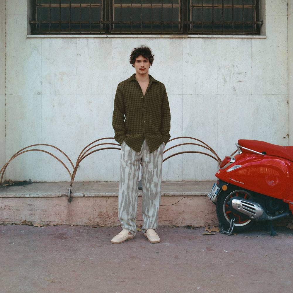 A cropped image of a man leaning against a dark wooden wall. He is wearing a light green and cream Terry cotton pants in a banana leaf design.