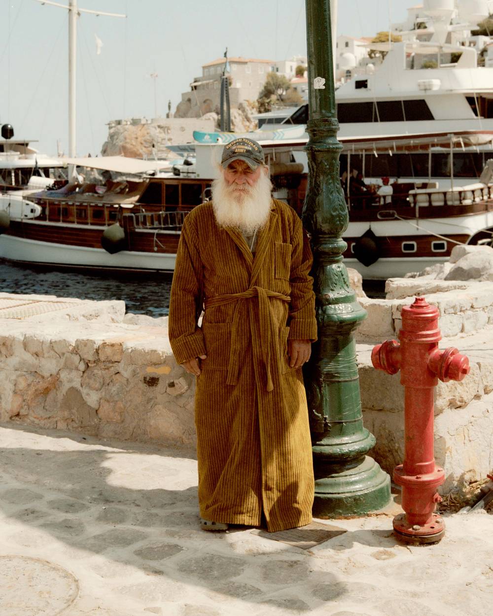 A horizontal picture shot from behind of two elderly men sitting on a bench. The man on the left is wearing a red and white paisley bathrobe and the man on the right a red and white wavy pattern bathrobe. The two men look out over a bay on the Mediterranean where people are swimming and enjoying the sun weather.