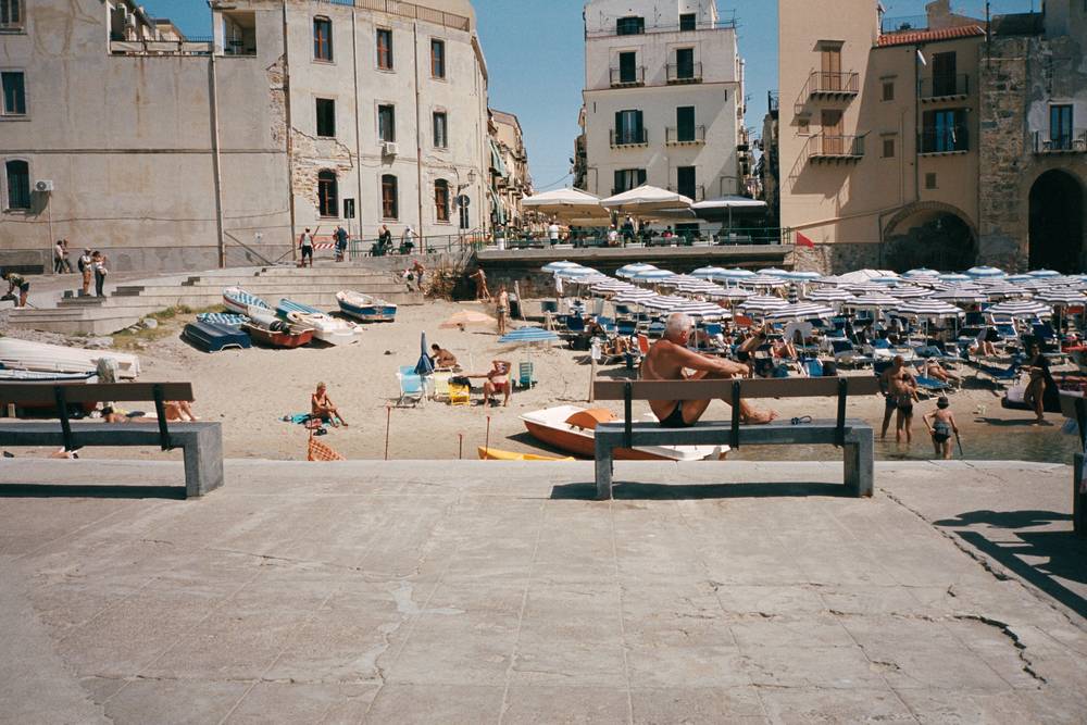 The image features a man sitting on a bench with his feet resting on it as well. In the background you can spot all the beachgoers, umbrellas and boats lined up on the beach. Behind the beach there's a lot of old buildings.