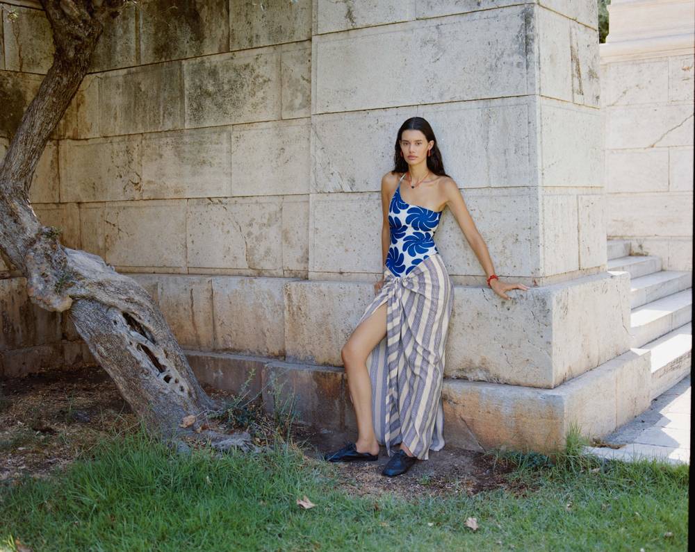 A woman in a blue and tan patterned top and a striped wrap skirt stands against a textured beige wall, conveying a minimalist and contemporary aesthetic.