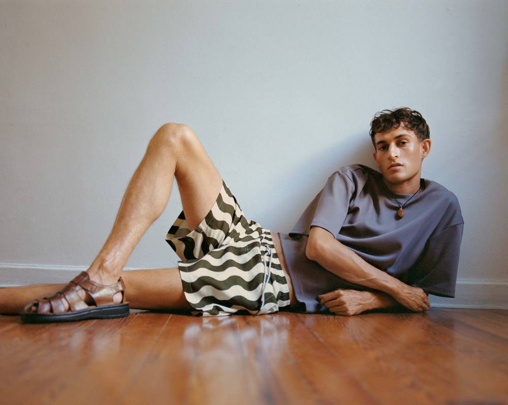 A cropped image of a young man wearing a pair of navy swim shorts with a yellow sun print. He has a burgundy and white printed towel thrown on his shoulder as well. Blurred in the front of the picture there is two men sitting one is wearing black and the other one is wearing a purple velour shirt.  