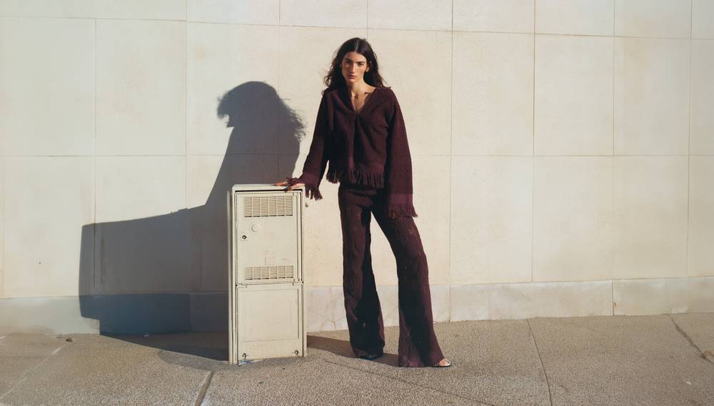 A woman standing next to an antique cupboard wearing a burgundy bikini top, a pair of white and burgundy patterned long pants and dark sandals. In the front of the picture there's a light wooden dining table and chairs.