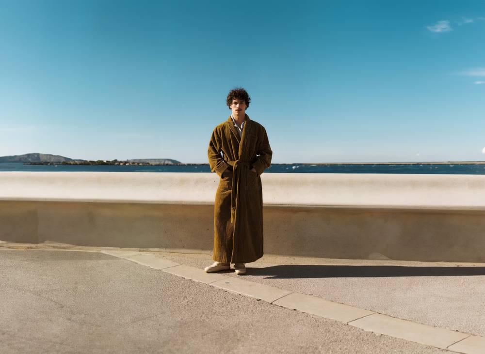 A horizontal picture shot from behind of two elderly men sitting on a bench. The man on the left is wearing a red and white paisley bathrobe and the man on the right a red and white wavy pattern bathrobe. The two men look out over a bay on the Mediterranean where people are swimming and enjoying the sun weather.