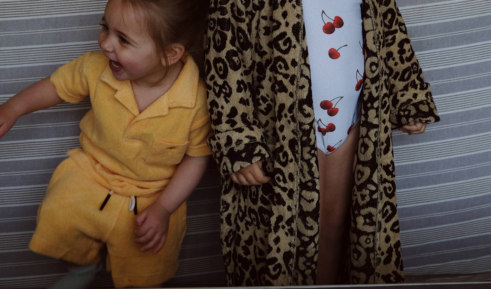 There are two kids, the one on the left is wearing a bright yellow Terry set, shorts and a shortsleeved shirt. The kid on the right is wearing a cherry printed bathing suit and a leopard printed robe on top. 