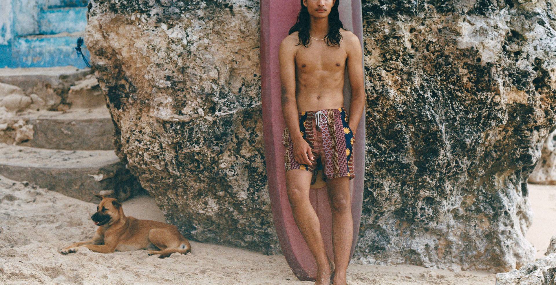 A cropped image of a young man wearing a pair of navy swim shorts with a yellow sun print. He has a burgundy and white printed towel thrown on his shoulder as well. Blurred in the front of the picture there is two men sitting one is wearing black and the other one is wearing a purple velour shirt.  