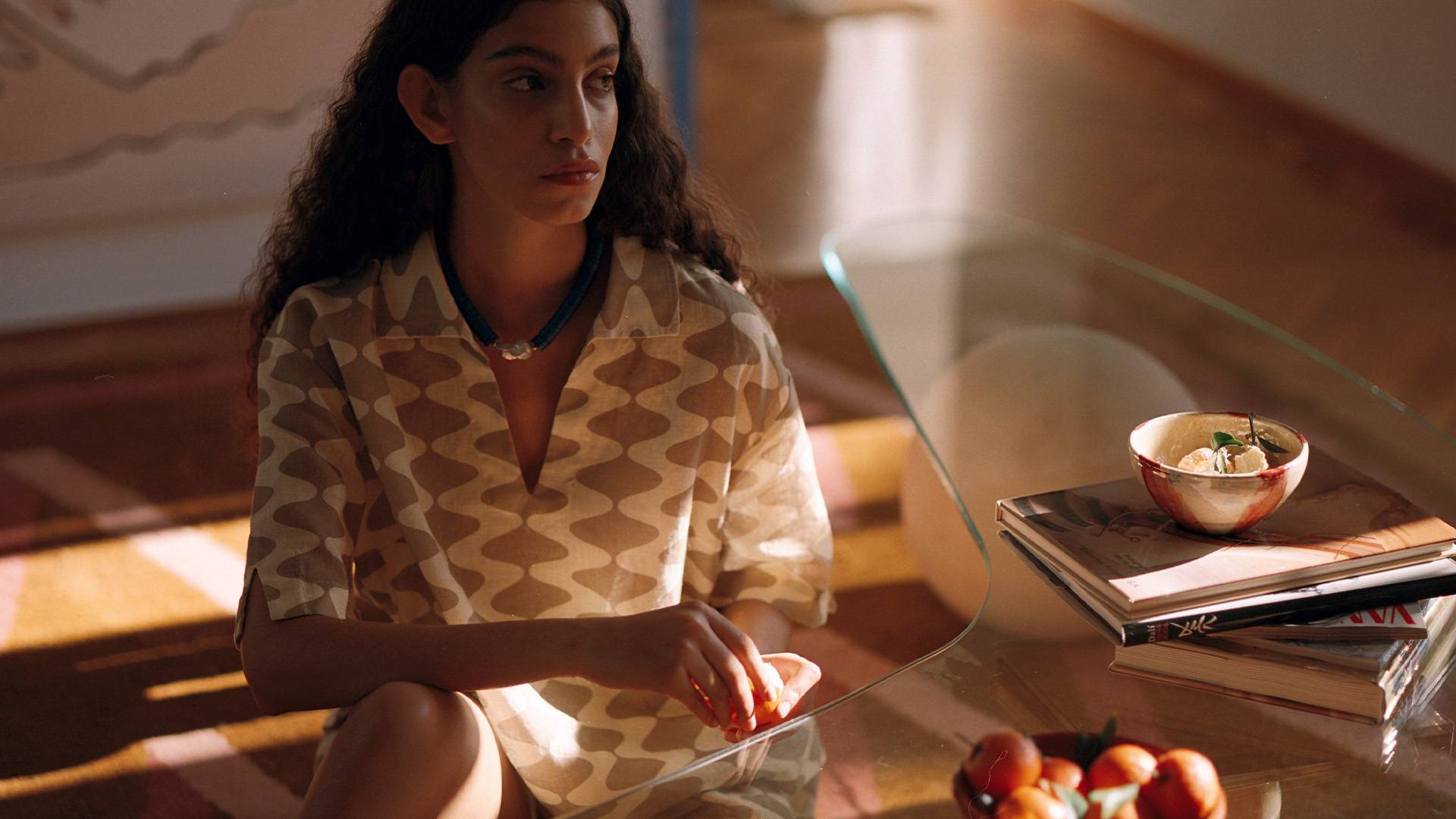 A woman sitting on the floor next to a glass-table with the light seeping in creating a shadows all around. She is wearing a cream and sand printed collared shirt and matching shorts.
