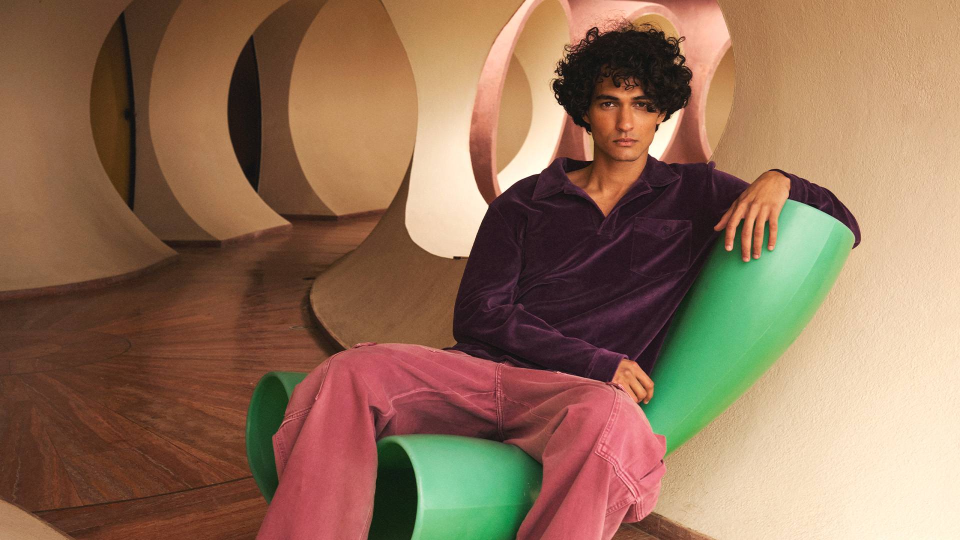 A dark and curly haired man is sitting on a bright green chair in a hallway. He is wearing a long sleeved deep purple velour shirt and washed red cargo pants.