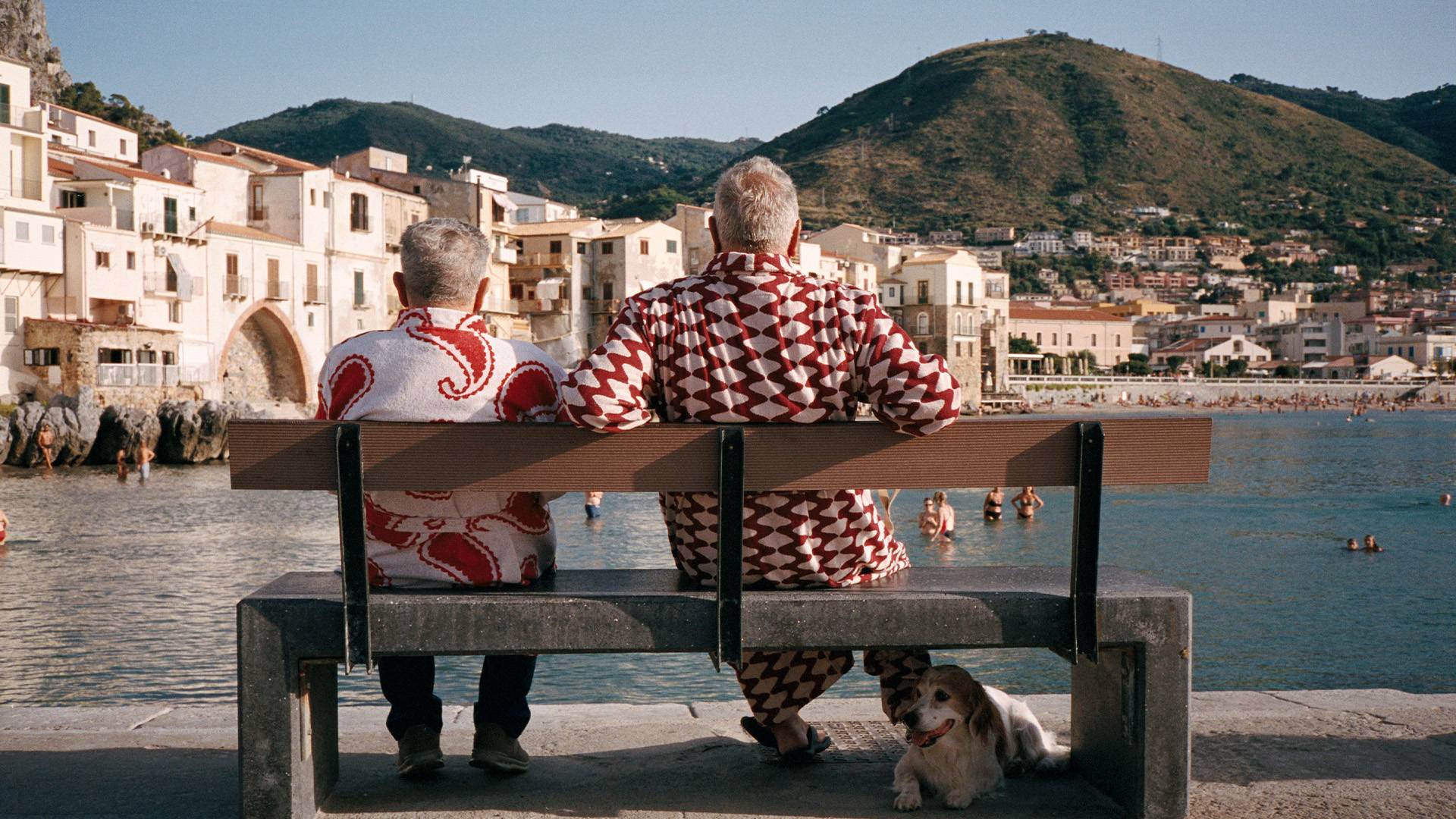 A horizontal picture shot from behind of two elderly men sitting on a bench. The man on the left is wearing a red and white paisley bathrobe and the man on the right a red and white wavy pattern bathrobe. The two men look out over a bay on the Mediterranean where people are swimming and enjoying the sun weather.