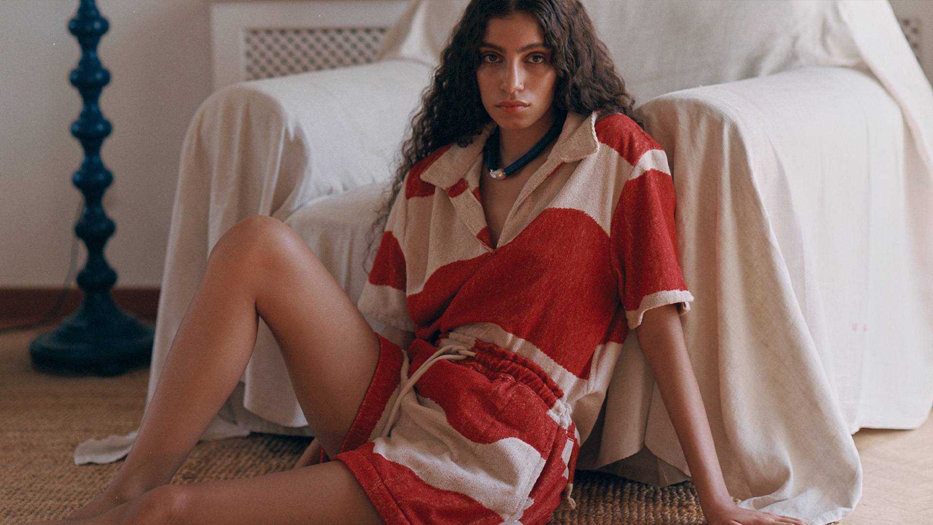 A woman sitting infront of a sheet-covered lounge chair. She is wearing a red and white printed pair of Terry shorts and a matching collared shirt.