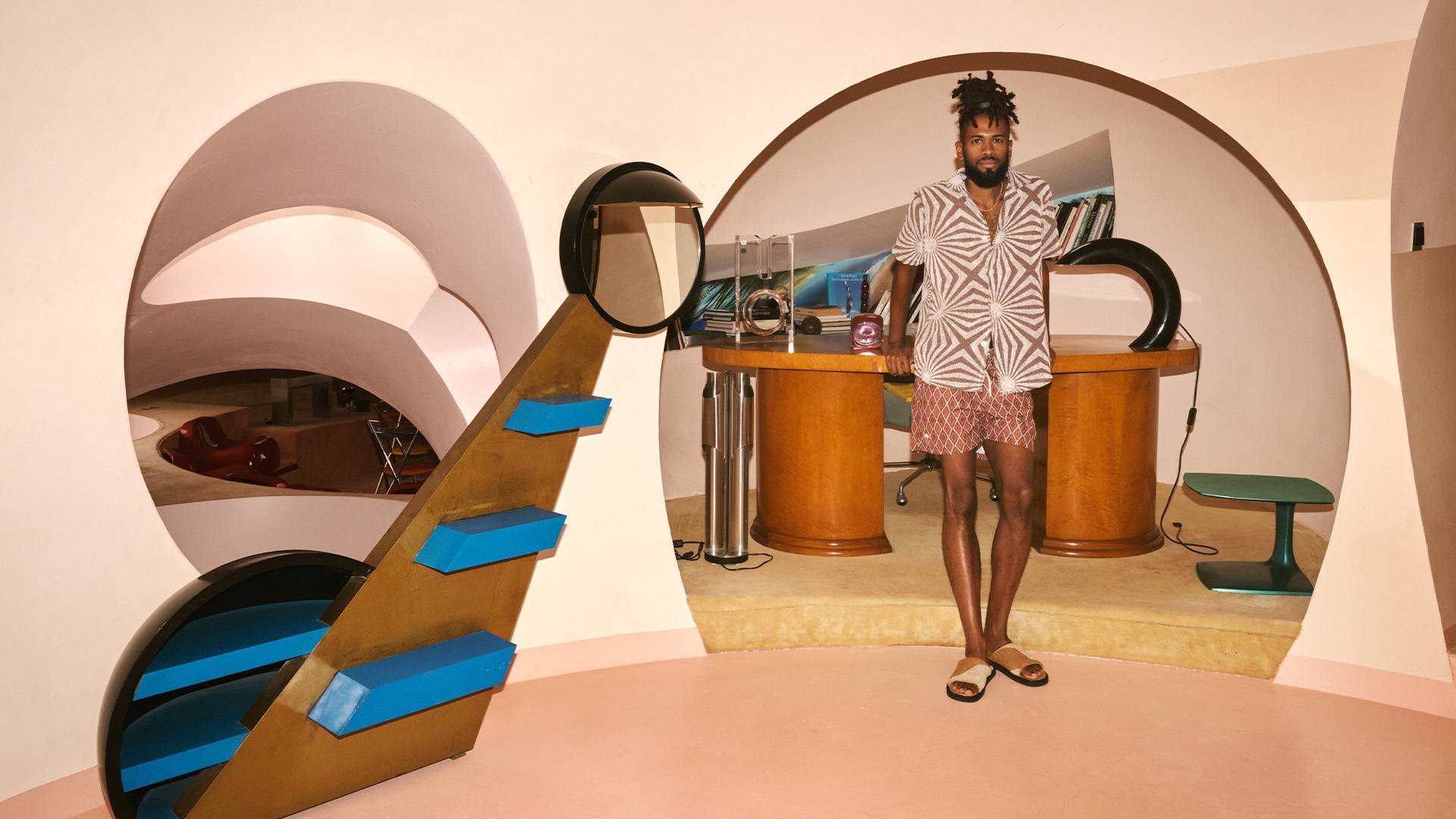 A man standing in an arched opening to an office, leaning on the desk. He is wearing a brown and white printed Terry shirt, a pair of rusty red diamond printed swim shorts and a pair of caramel brown and cram printed Terry slippers. The room he's standing in is a light shade of pink.