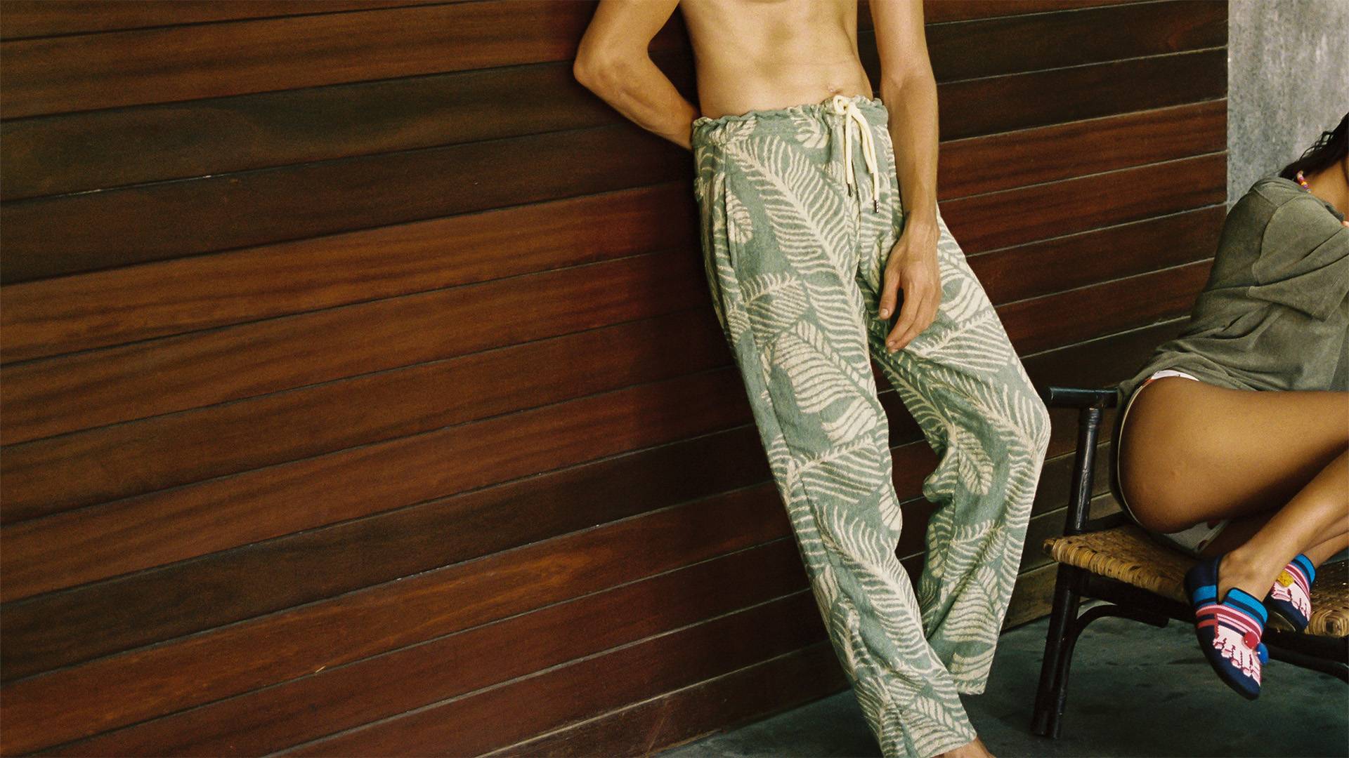 A cropped image of a man leaning against a dark wooden wall. He is wearing a light green and cream Terry cotton pants in a banana leaf design.