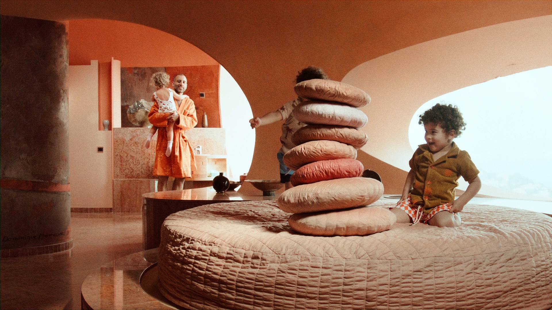 Two kids sitting on a round bed playing and building a tower with orange pillows. They are wearing Terry shirts with palmtrees on and printed swim shorts. A man is standing in the background wearing a orange robe holding another kid wearing a cherry printed swimsuit.