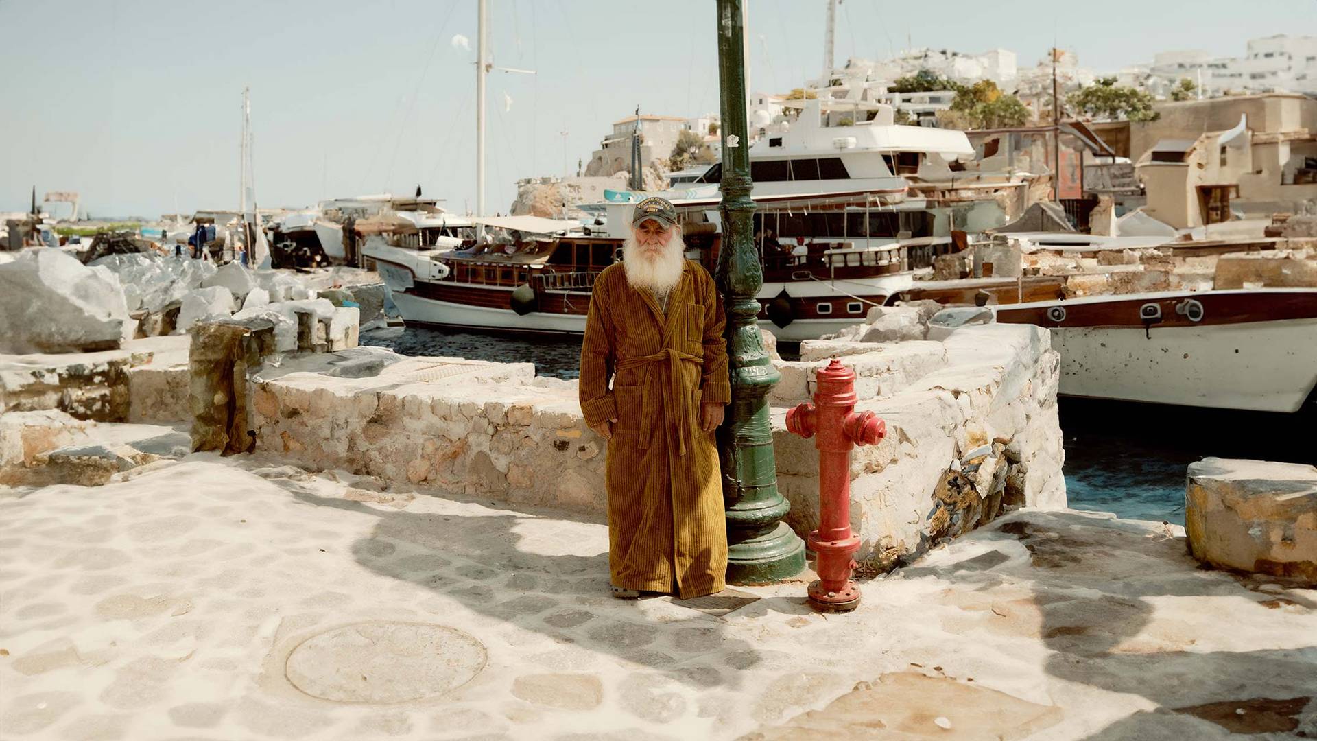 A horizontal picture shot from behind of two elderly men sitting on a bench. The man on the left is wearing a red and white paisley bathrobe and the man on the right a red and white wavy pattern bathrobe. The two men look out over a bay on the Mediterranean where people are swimming and enjoying the sun weather.