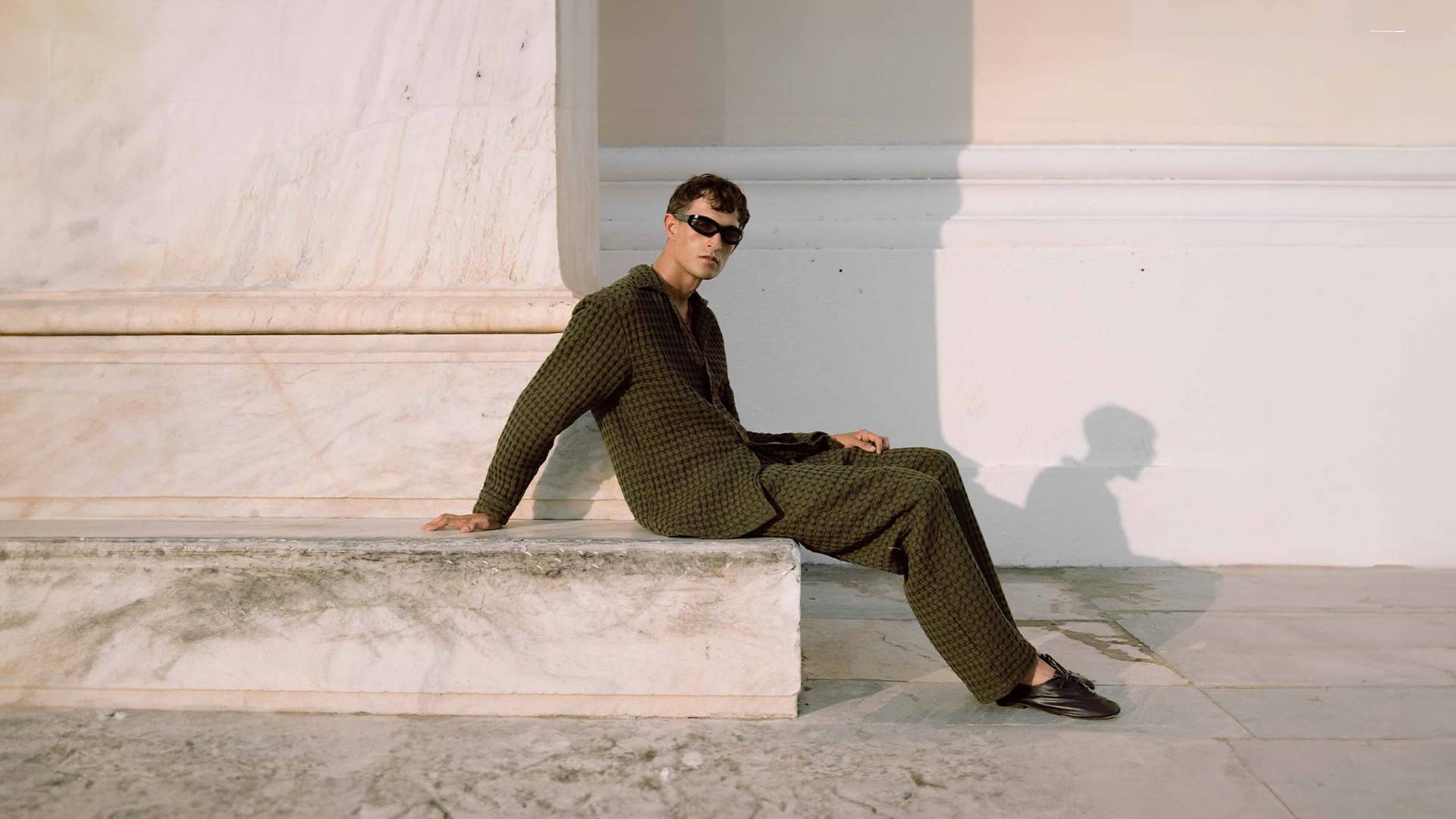 A cropped image of a man leaning against a dark wooden wall. He is wearing a light green and cream Terry cotton pants in a banana leaf design.