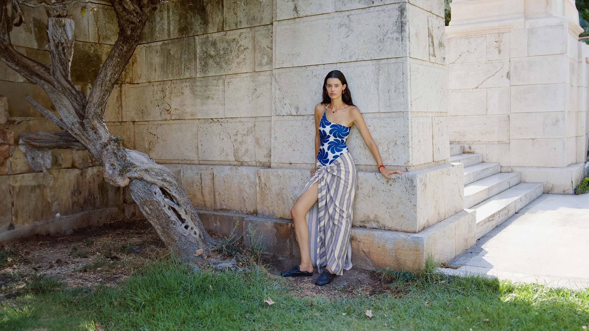 A woman in a blue and tan patterned top and a striped wrap skirt stands against a textured beige wall, conveying a minimalist and contemporary aesthetic.