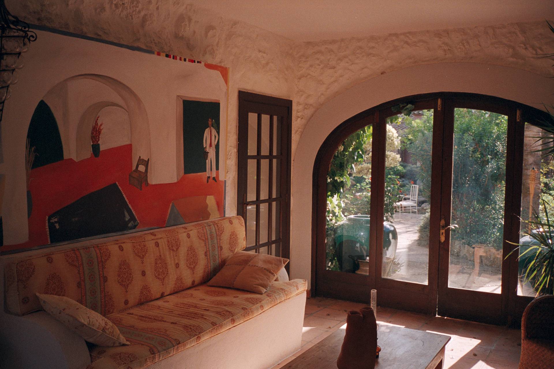 View of living room in Case Calandra, Mallorca. The image features a sofa with yellow and orange cushions, an arched glass doorway leading out to the garden and a huge painting with red and white motifs.