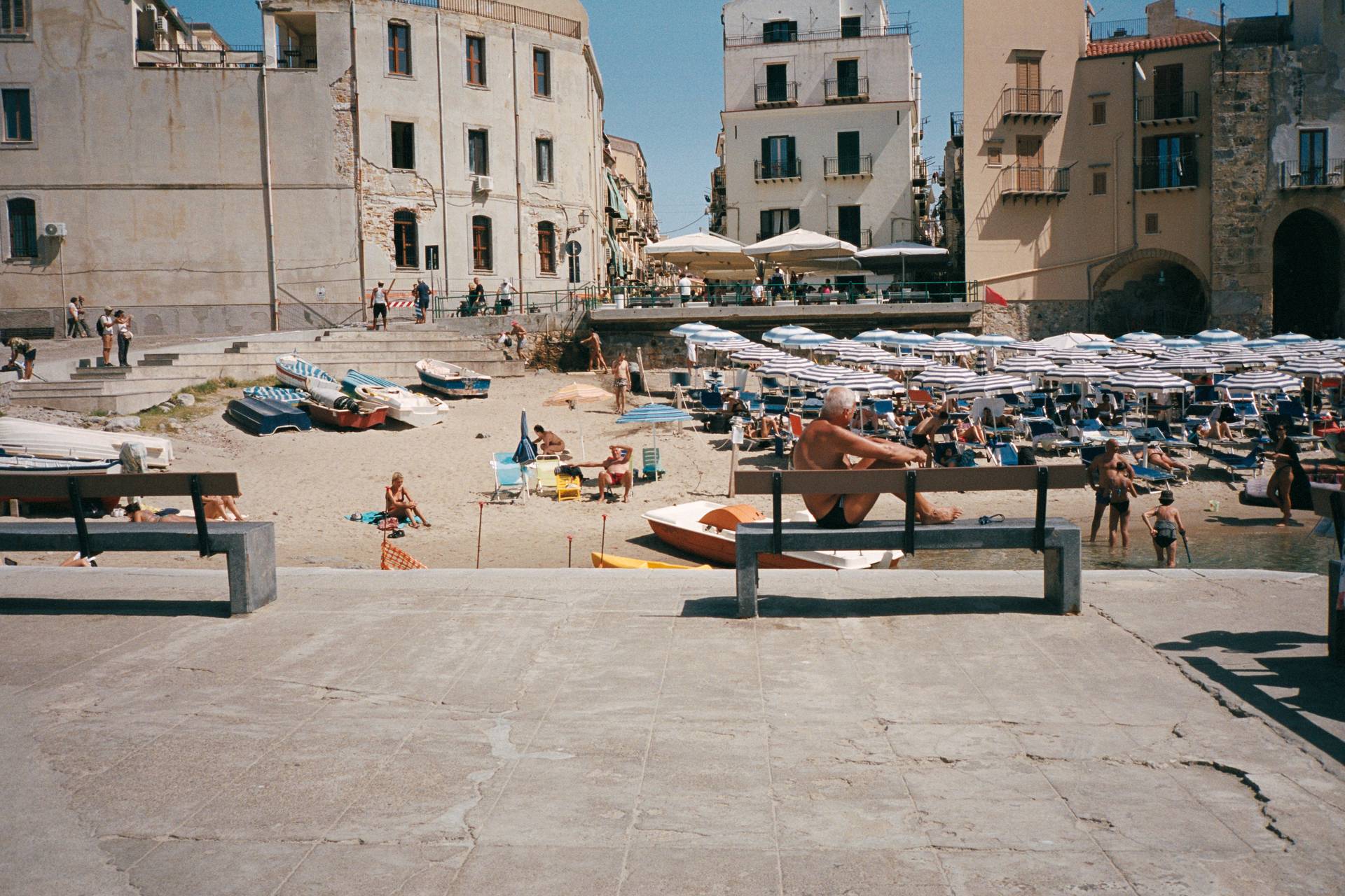 The image features a man sitting on a bench with his feet resting on it as well. In the background you can spot all the beachgoers, umbrellas and boats lined up on the beach. Behind the beach there's a lot of old buildings.