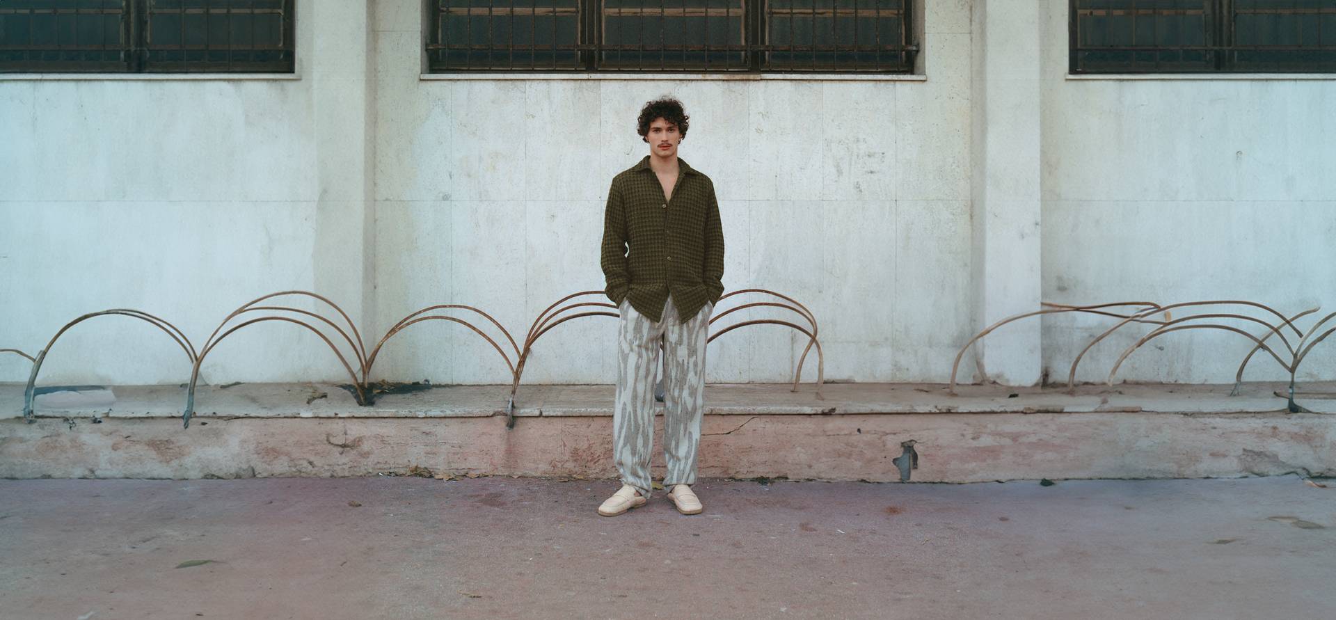 A cropped image of a man leaning against a dark wooden wall. He is wearing a light green and cream Terry cotton pants in a banana leaf design.