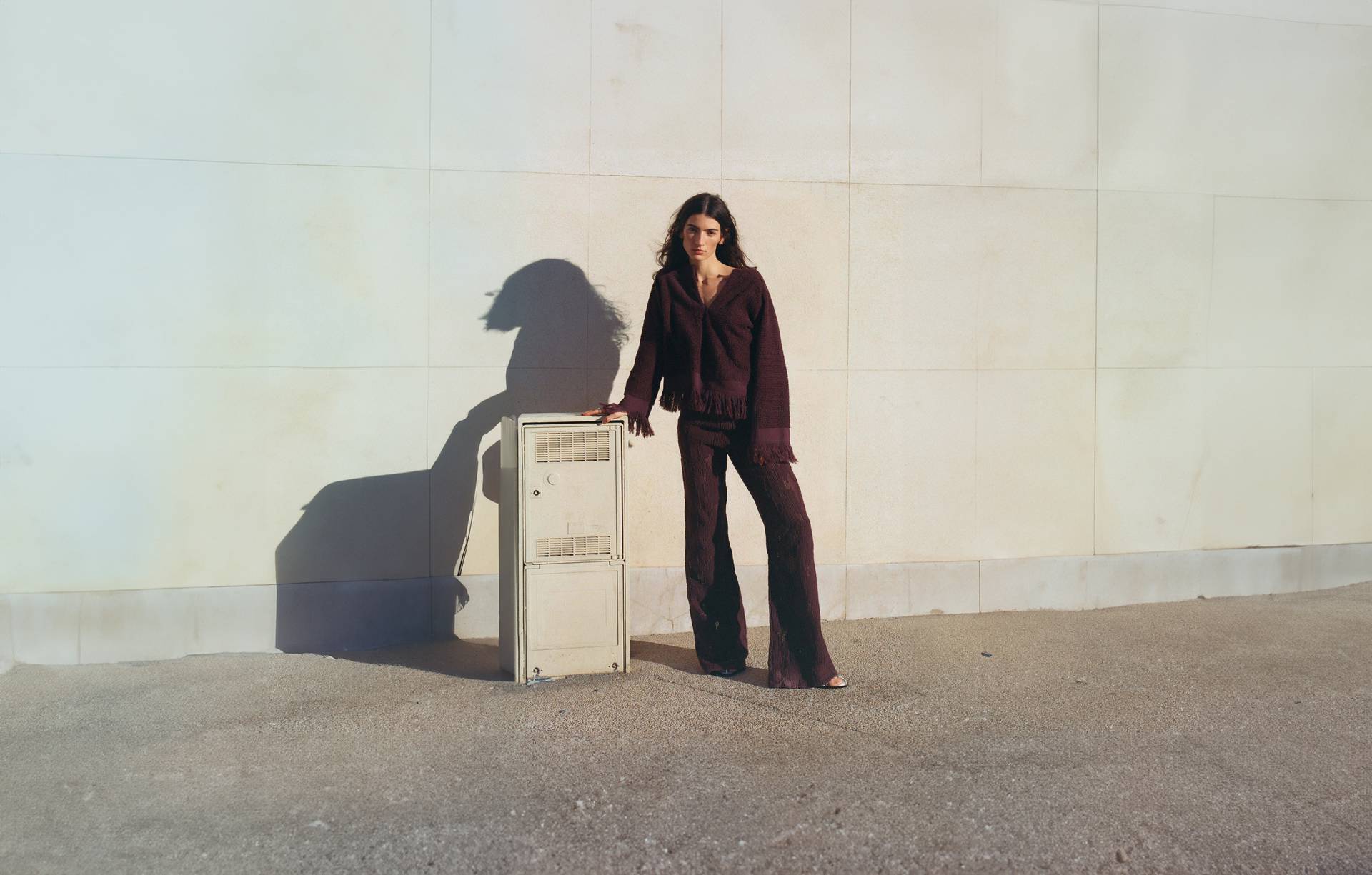 A woman standing next to an antique cupboard wearing a burgundy bikini top, a pair of white and burgundy patterned long pants and dark sandals. In the front of the picture there's a light wooden dining table and chairs.