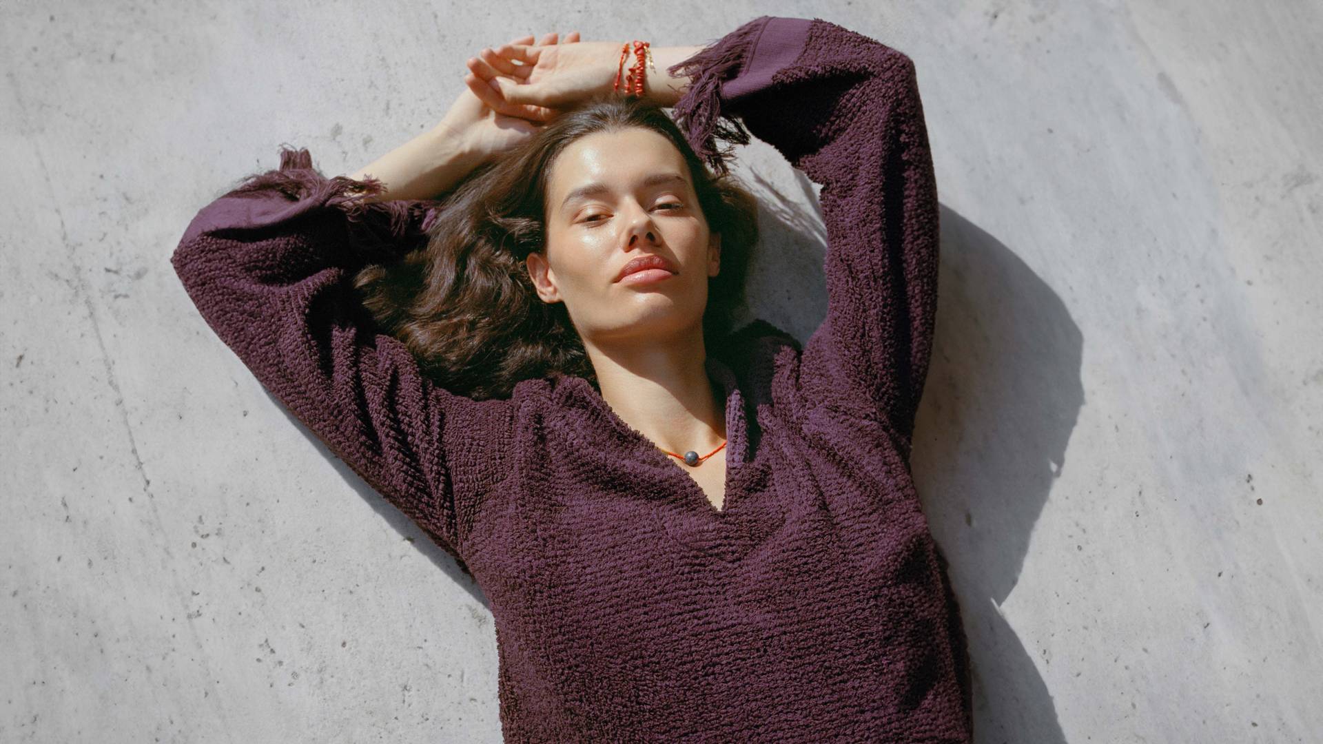 A woman sitting on the floor next to a glass-table with the light seeping in creating a shadows all around. She is wearing a cream and sand printed collared shirt and matching shorts.