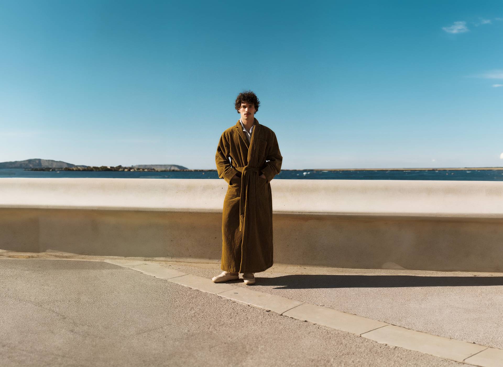 A horizontal picture shot from behind of two elderly men sitting on a bench. The man on the left is wearing a red and white paisley bathrobe and the man on the right a red and white wavy pattern bathrobe. The two men look out over a bay on the Mediterranean where people are swimming and enjoying the sun weather.