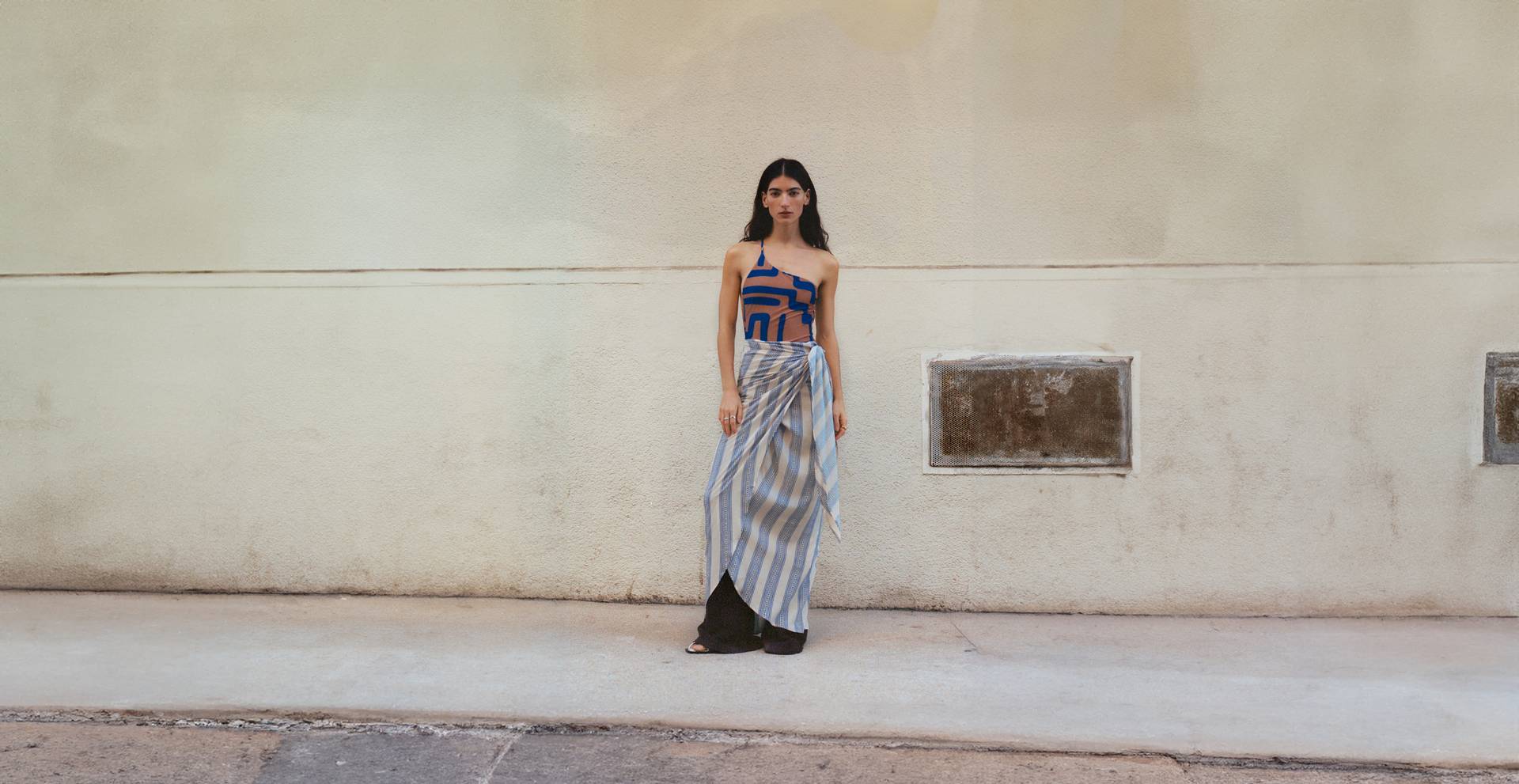 A woman in a blue and tan patterned top and a striped wrap skirt stands against a textured beige wall, conveying a minimalist and contemporary aesthetic.