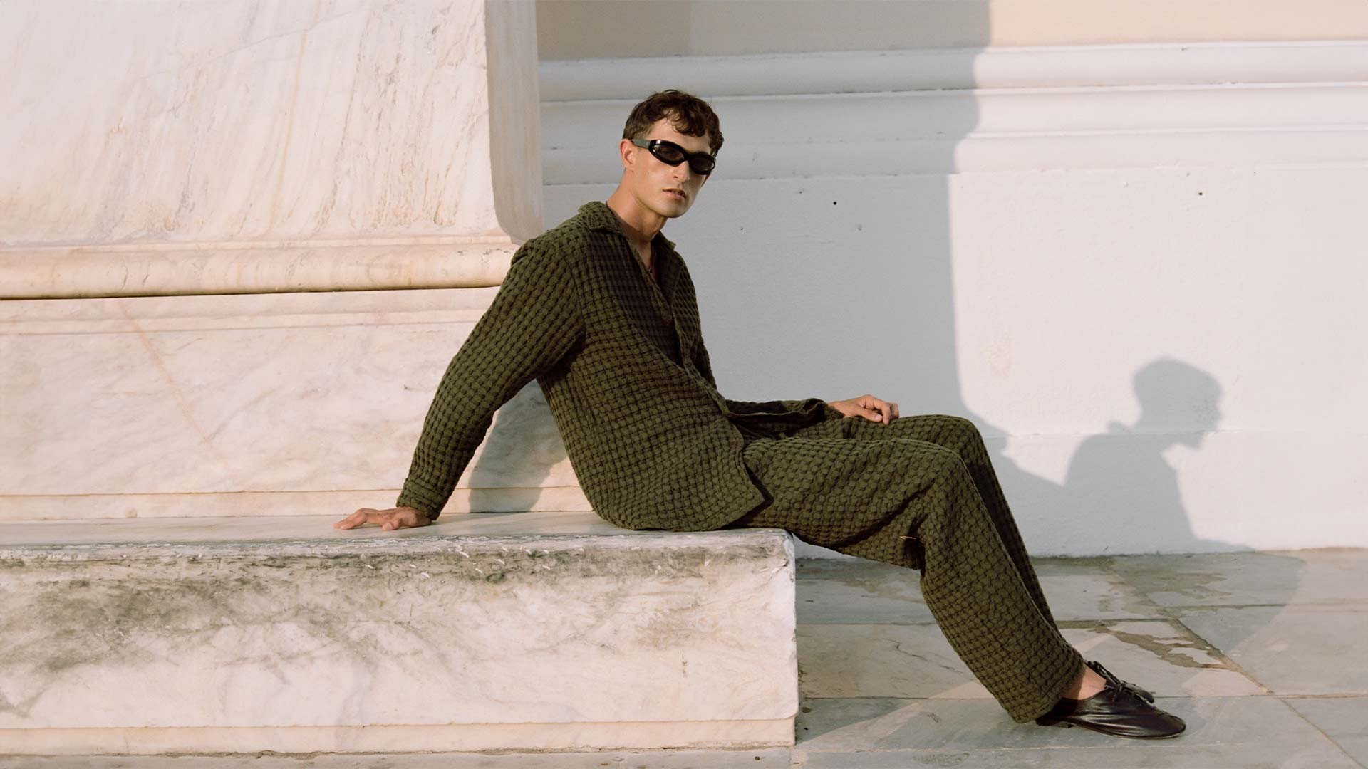 Model wearing a stylish green houndstooth-patterned outfit, consisting of a relaxed-fit shirt and matching trousers, standing confidently against a white marble monument. The model is accessorized with dark sunglasses