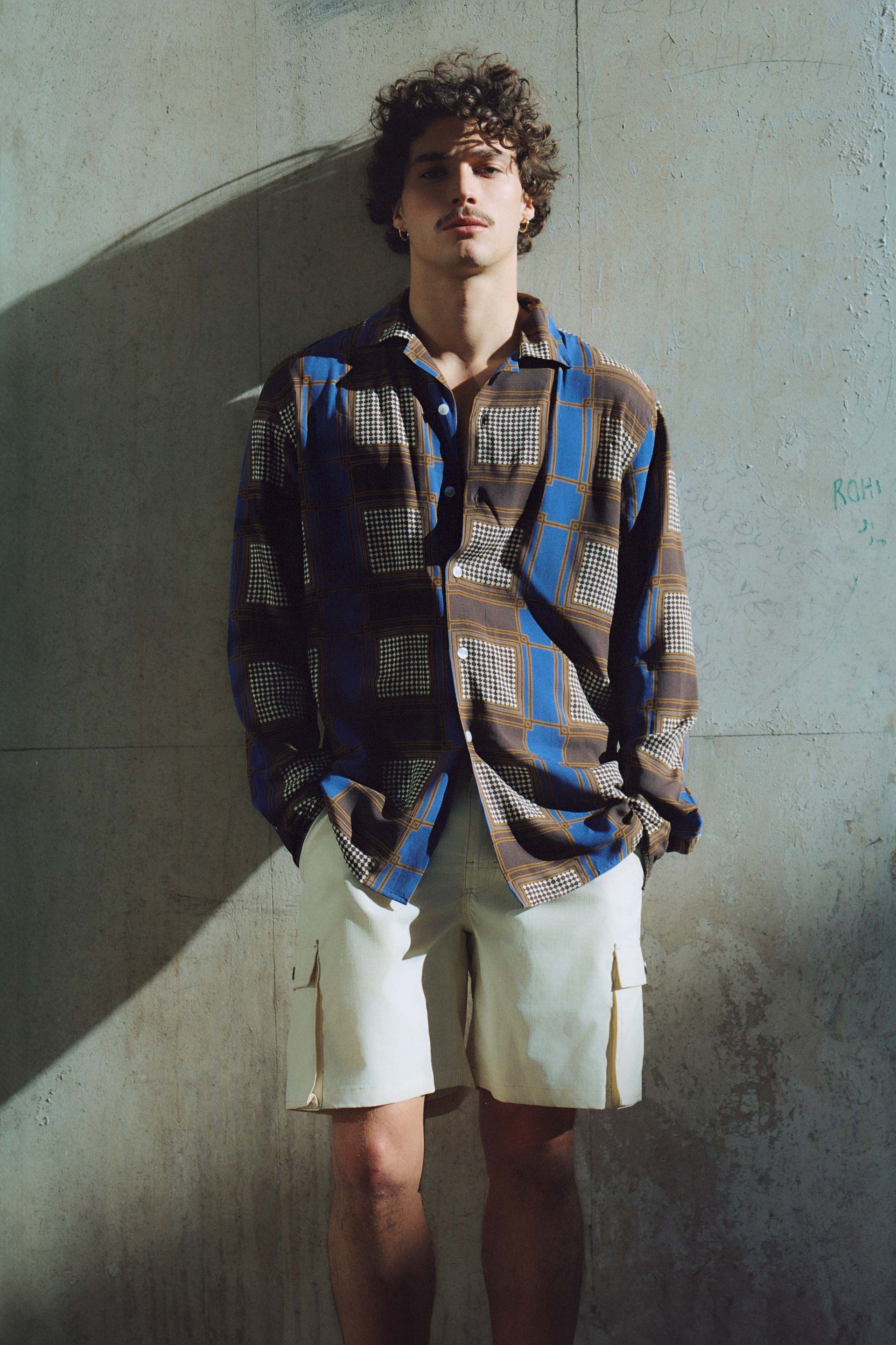 A man wearing a patterned shirt and light-colored shorts stands against a textured wall. Sunlight and shadows add depth to the urban setting, highlighting his casual yet stylish look.