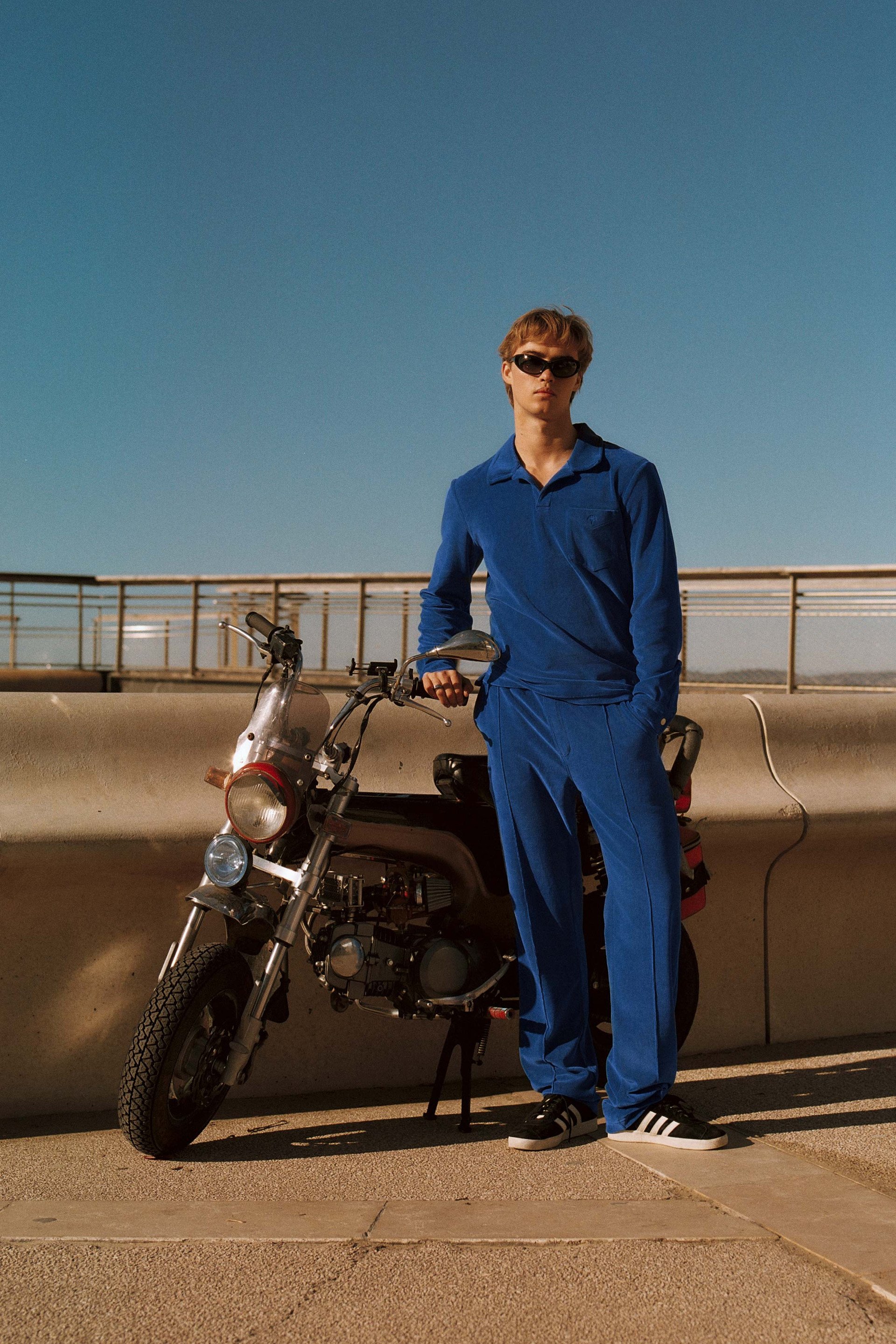 A young man in a blue tracksuit and sunglasses stands next to a small motorcycle against a clear blue sky, with a modern concrete and metal backdrop.