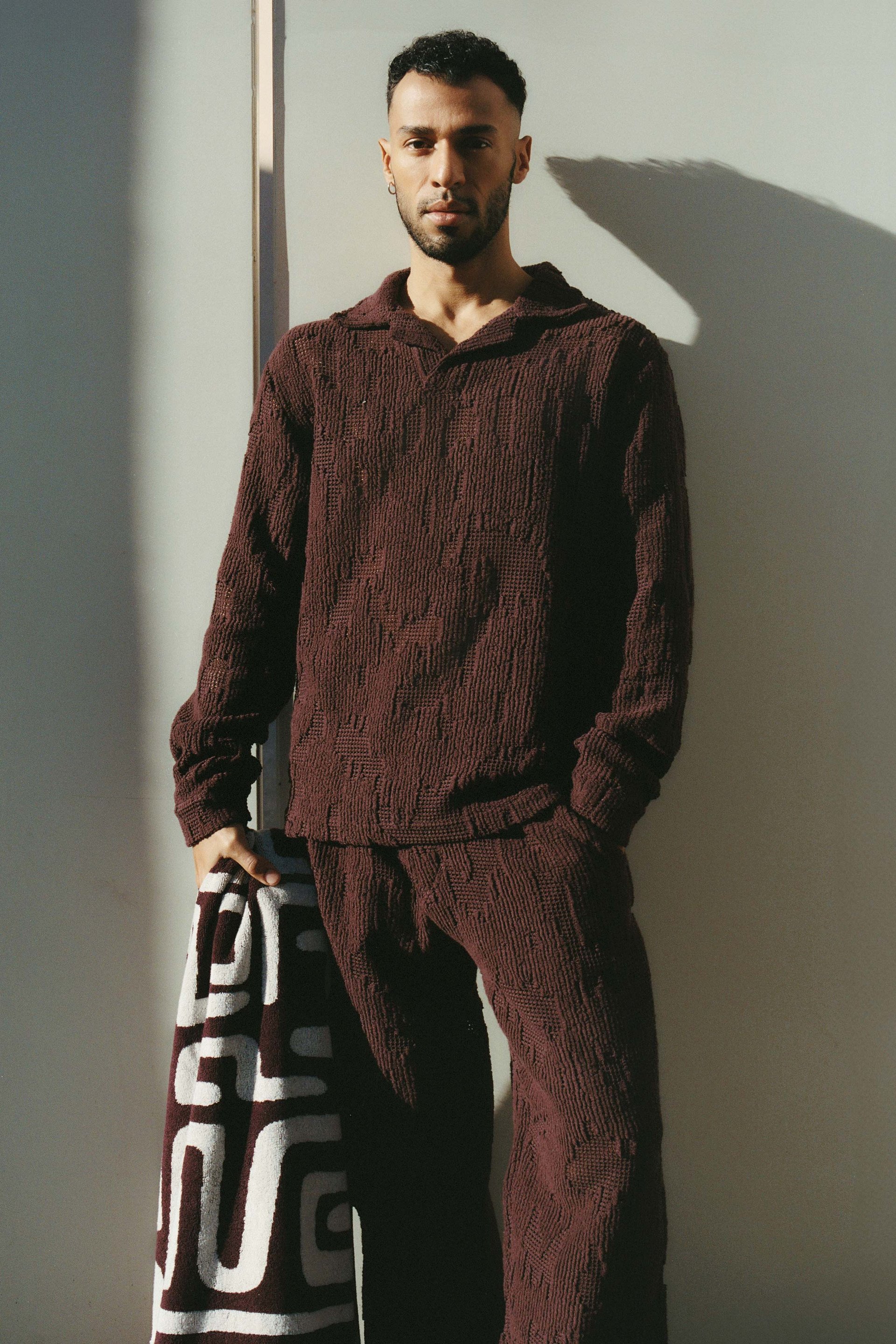 A man in a maroon textured outfit stands against a light-colored wall, holding a patterned towel. The scene is illuminated by sunlight casting geometric shadows.