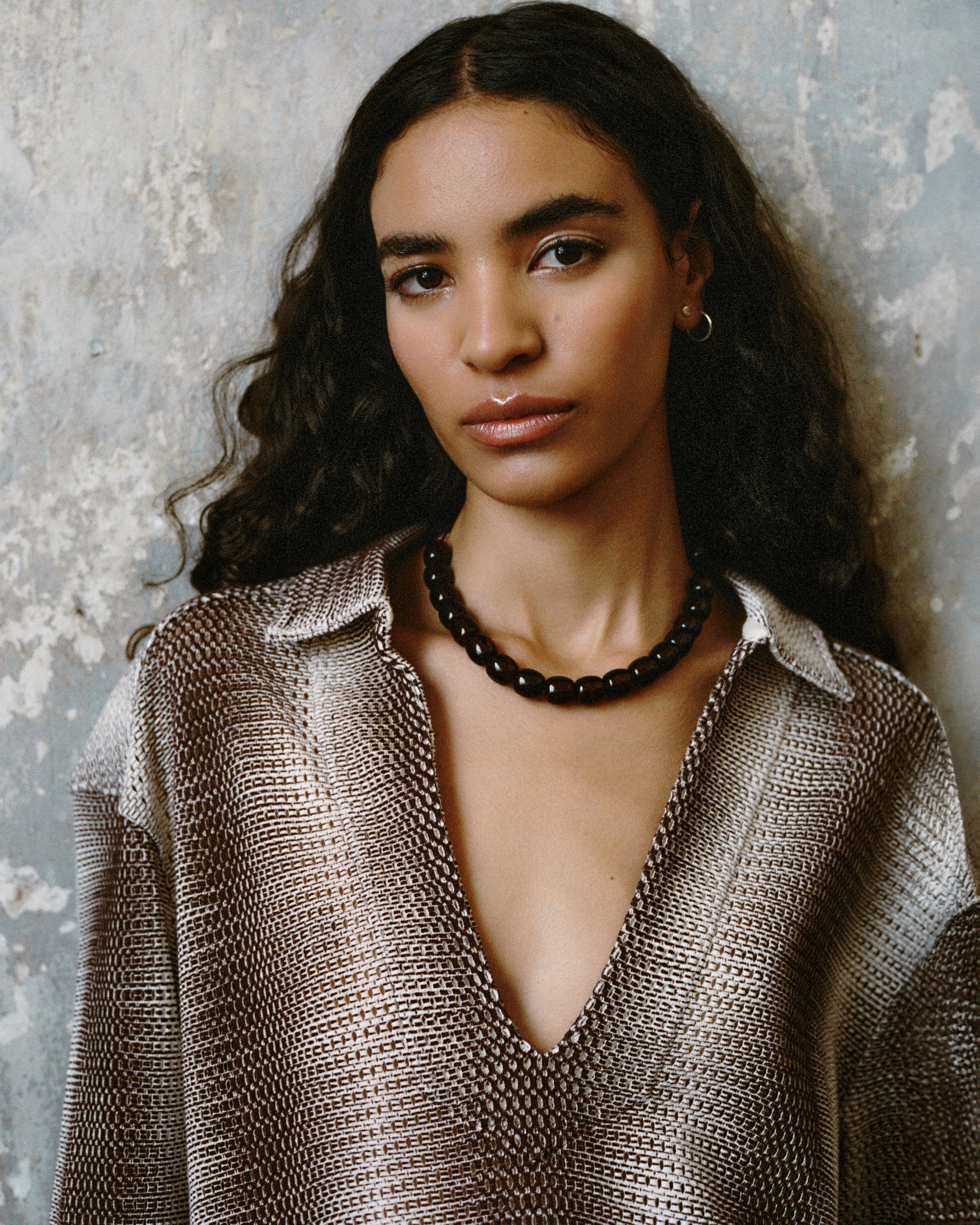 Model in a gradient knit dress with a deep neckline, paired with a black beaded necklace against a textured wall