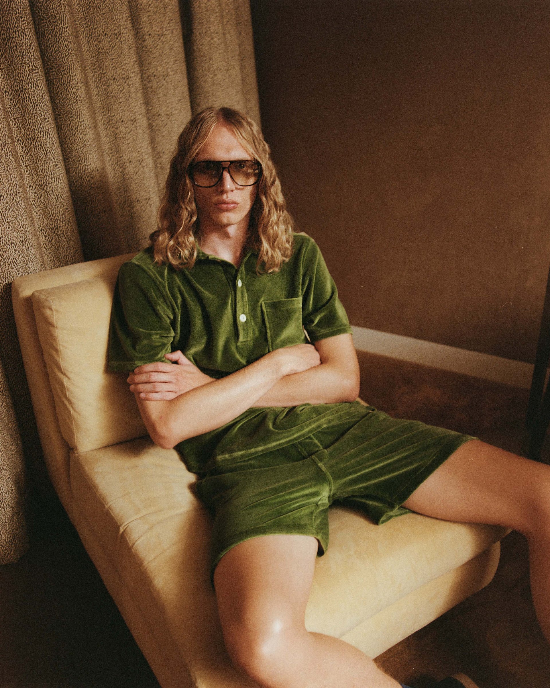 A model dressed in green velour shorts and polo, reclining on a beige chair in a retro-inspired interior, showcasing bold casual luxury.