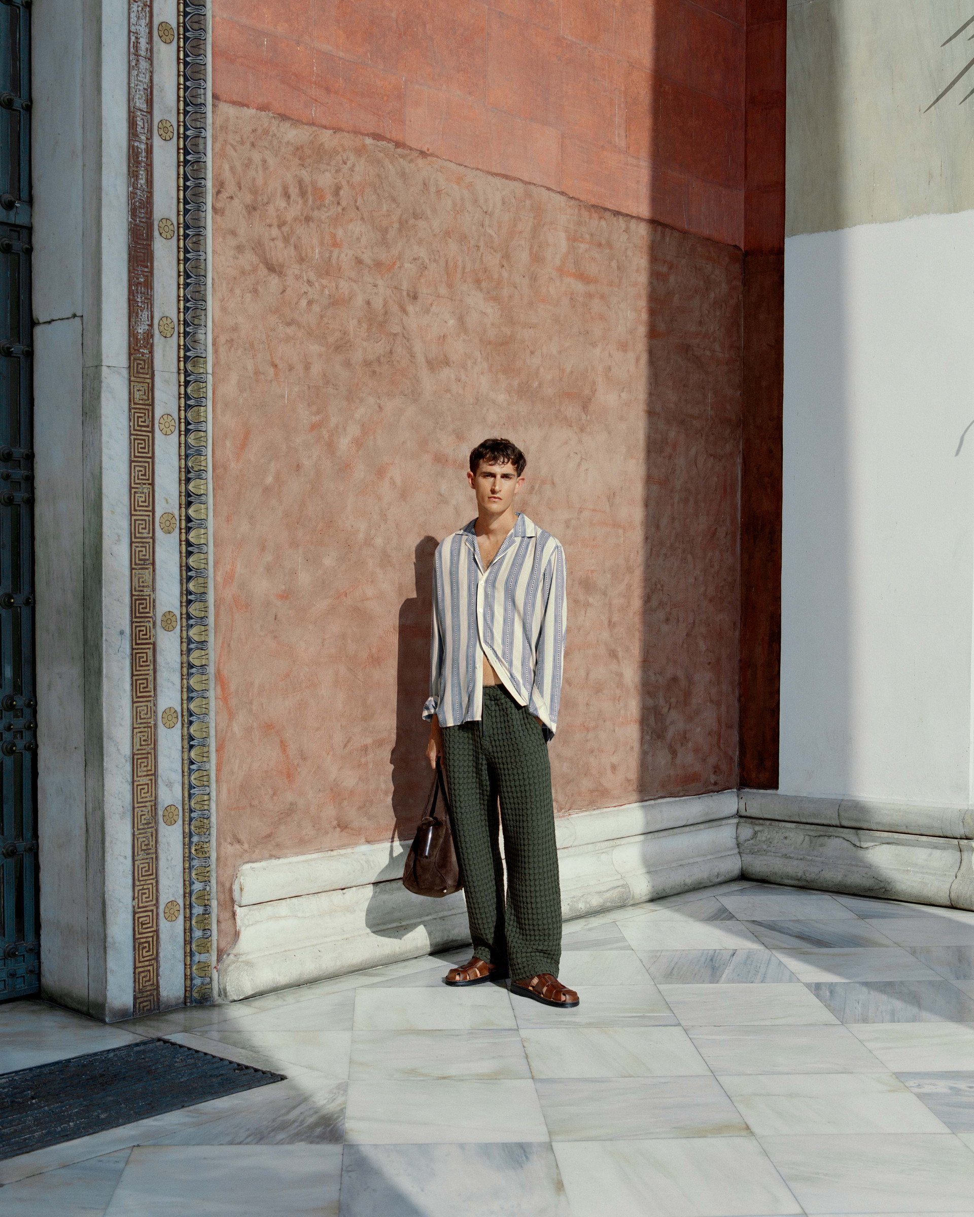 A man wearing a patterned shirt and light-colored shorts stands against a textured wall. Sunlight and shadows add depth to the urban setting, highlighting his casual yet stylish look.