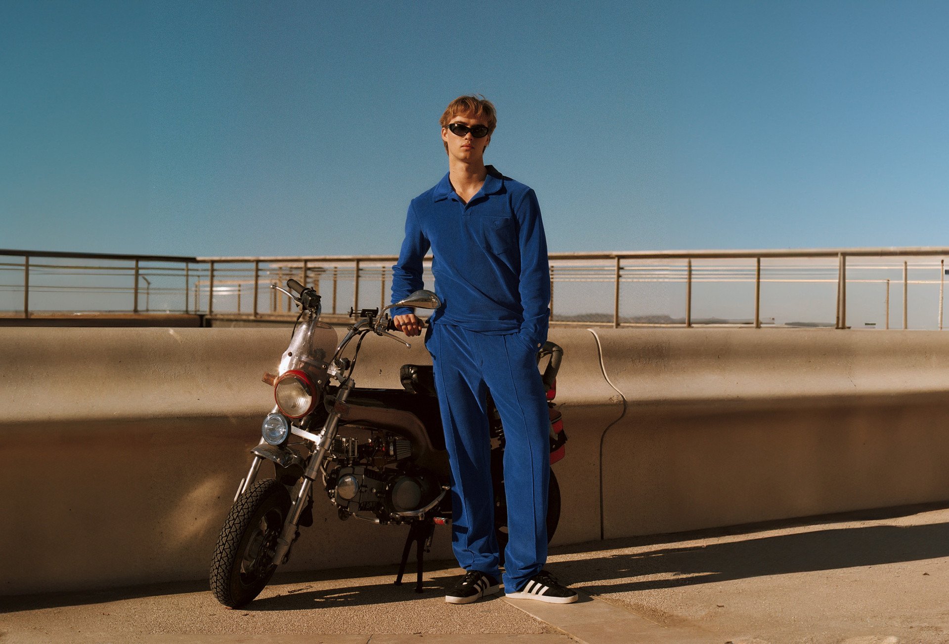 A young man in a blue velour set and sunglasses stands next to a small motorcycle against a clear blue sky, with a modern concrete and metal backdrop.