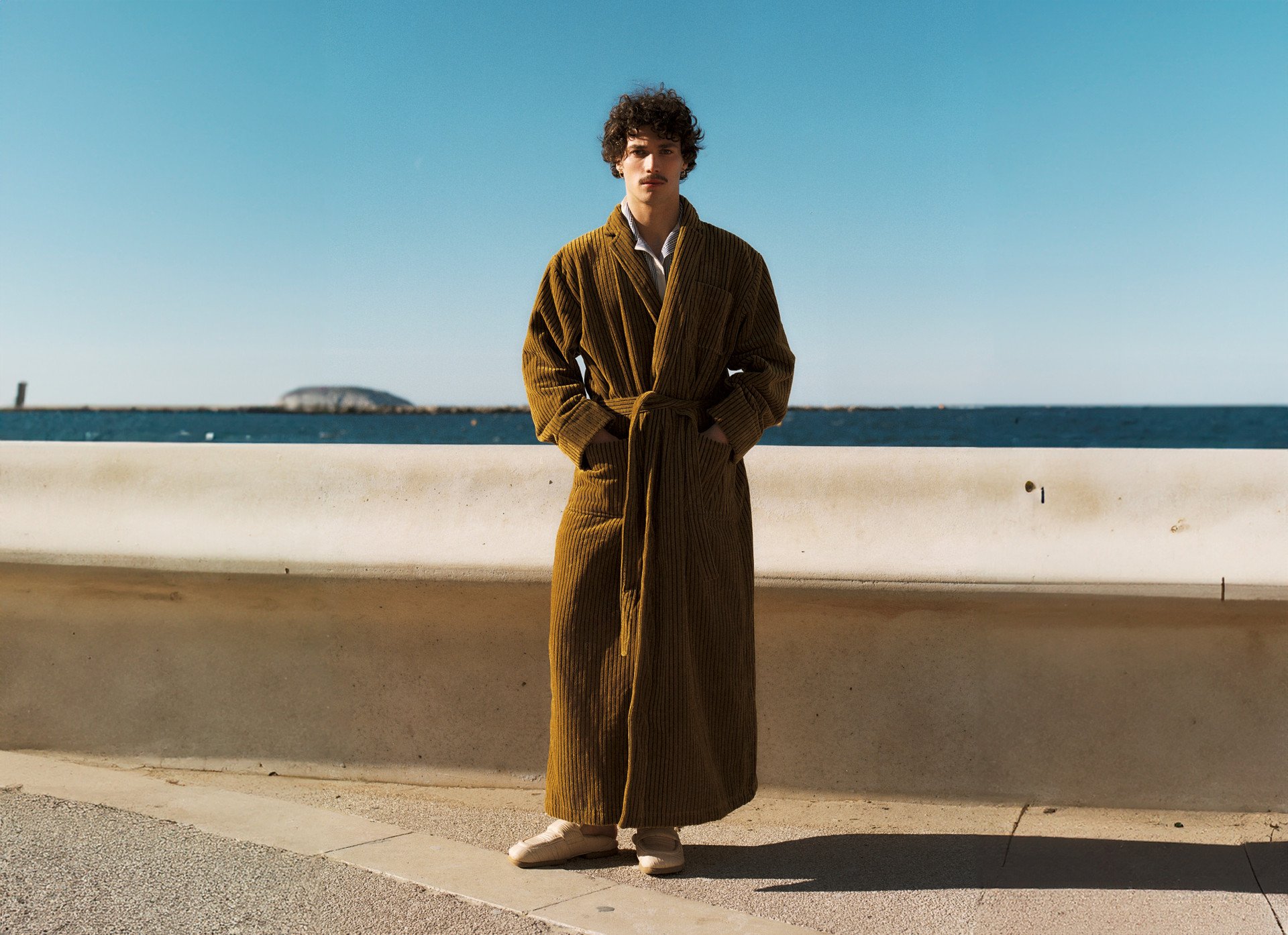 A man in a long, mustard-colored robe stands in front of a concrete barrier with a clear blue sky and ocean in the background, exuding a relaxed and stylish vibe.