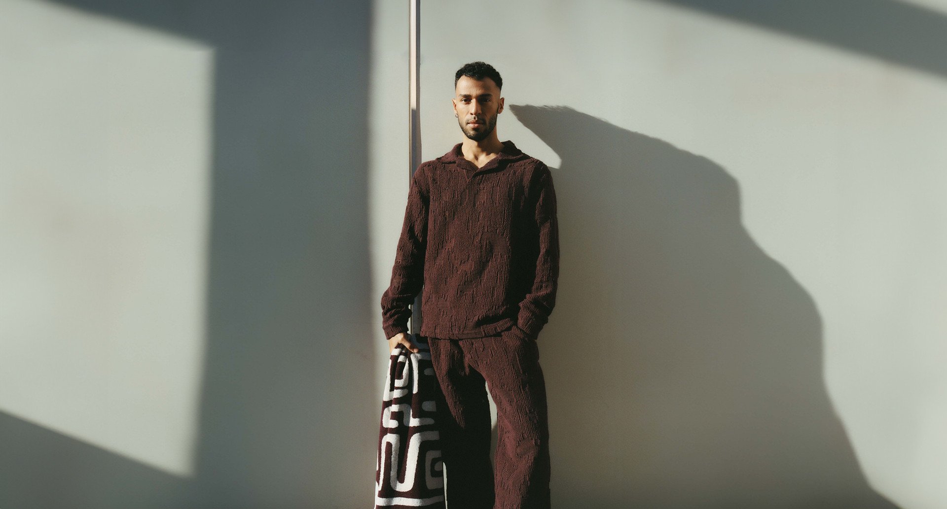 A man in a maroon textured outfit stands against a light-colored wall, holding a patterned towel. The scene is illuminated by sunlight casting geometric shadows.